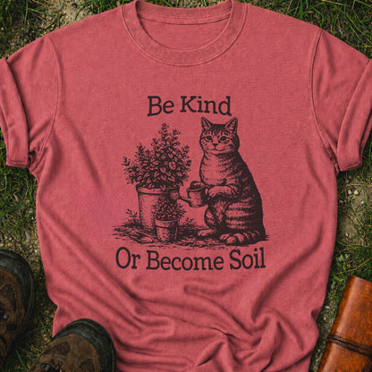 Dark Pink  t-shirt lying on a grassy surface.  the graphic is a Funny vintage-style gardening cat design featuring a cat watering plants with the phrase “Be Kind Or Become Soil.”