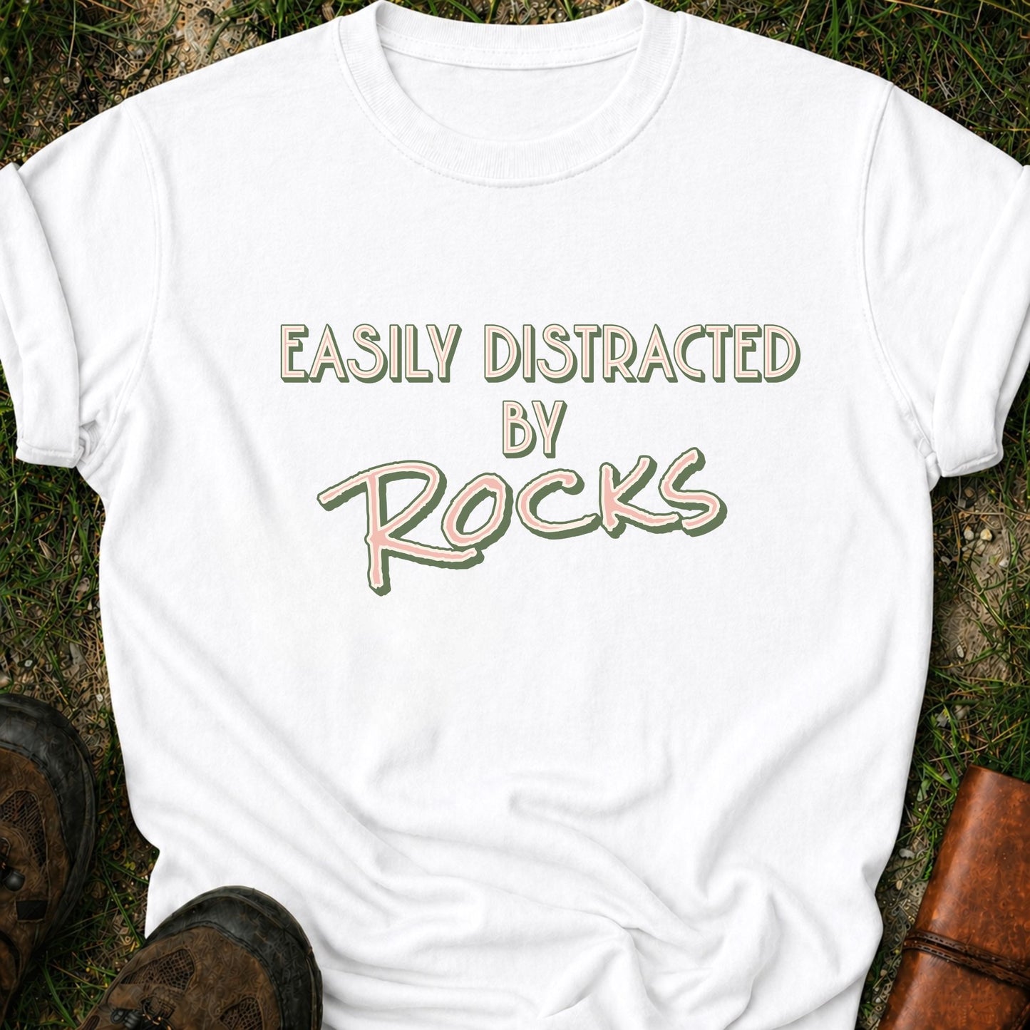 A white Retro text-based tee featuring the phrase “Easily Distracted by Rocks” in bold vintage lettering.  It is lying flat on a grassy surface.