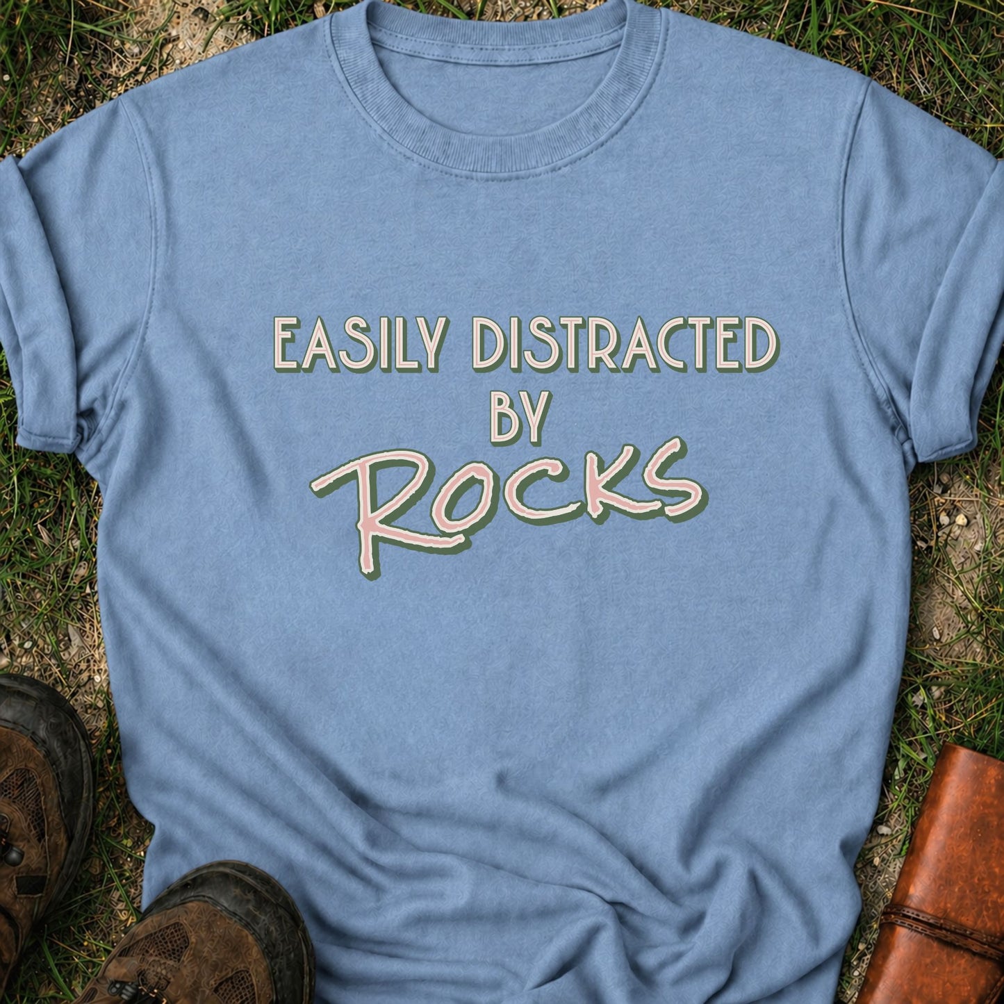 Blue Retro text-based tee featuring the phrase “Easily Distracted by Rocks” in bold vintage lettering.  It is lying flat on a grassy surface.