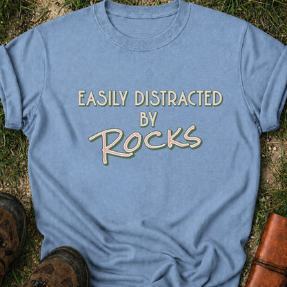 Blue Retro text-based tee featuring the phrase “Easily Distracted by Rocks” in bold vintage lettering.  It is lying flat on a grassy surface.