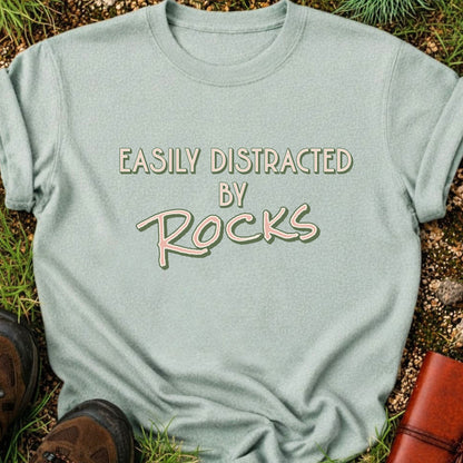 Light Green Retro text-based tee featuring the phrase “Easily Distracted by Rocks” in bold vintage lettering.  It is lying flat on a grassy surface.