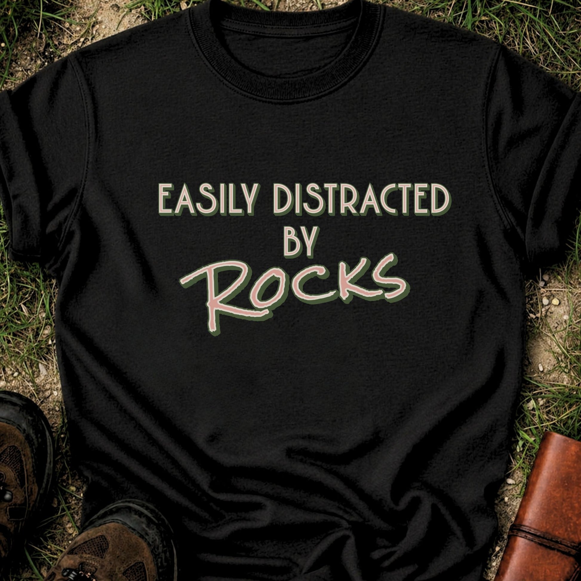 Black Retro text-based tee featuring the phrase “Easily Distracted by Rocks” in bold vintage lettering.  It is lying flat on a grassy surface.