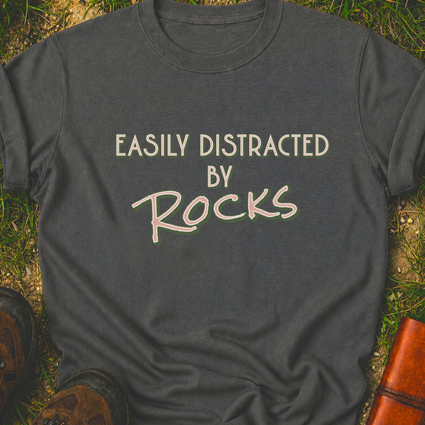 Garey Retro text-based tee featuring the phrase “Easily Distracted by Rocks” in bold vintage lettering.  It is lying flat on a grassy surface.