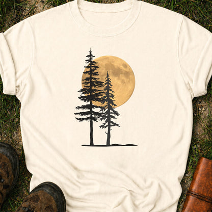 Ivory Tshirt with a graphic of  silhouetted pine trees against a glowing full moon. The Shirt is lying flat on a grassy surface.