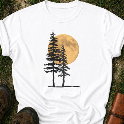 White Tshirt with a graphic of  silhouetted pine trees against a glowing full moon. The Shirt is lying flat on a grassy surface.