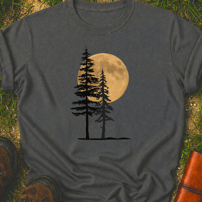 Dark Grey Tshirt with a graphic of  silhouetted pine trees against a glowing full moon. The Shirt is lying flat on a grassy surface.