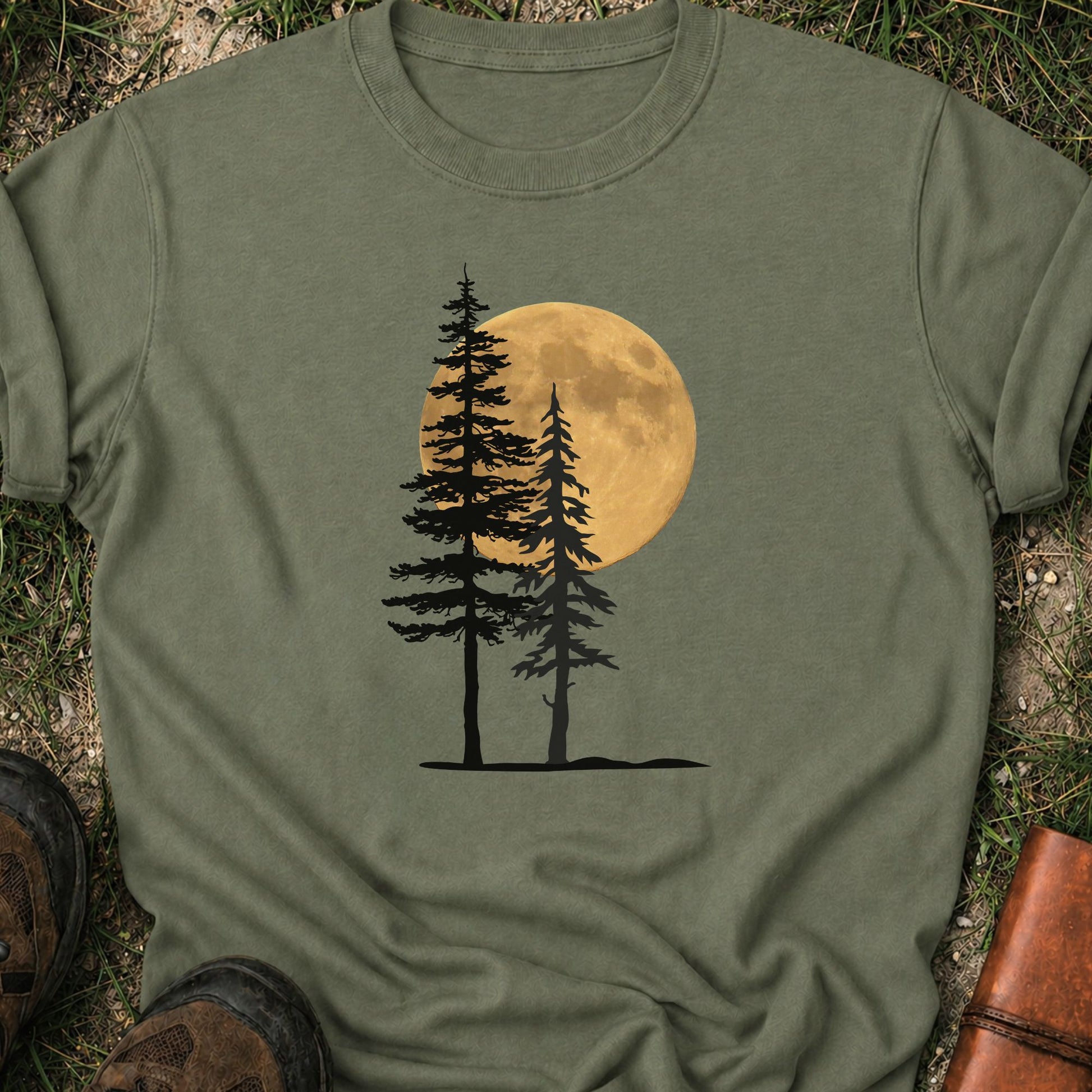 Moss Green Tshirt with a graphic of  silhouetted pine trees against a glowing full moon. The Shirt is lying flat on a grassy surface.