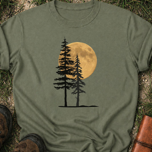 Moss Green Tshirt with a graphic of  silhouetted pine trees against a glowing full moon. The Shirt is lying flat on a grassy surface.