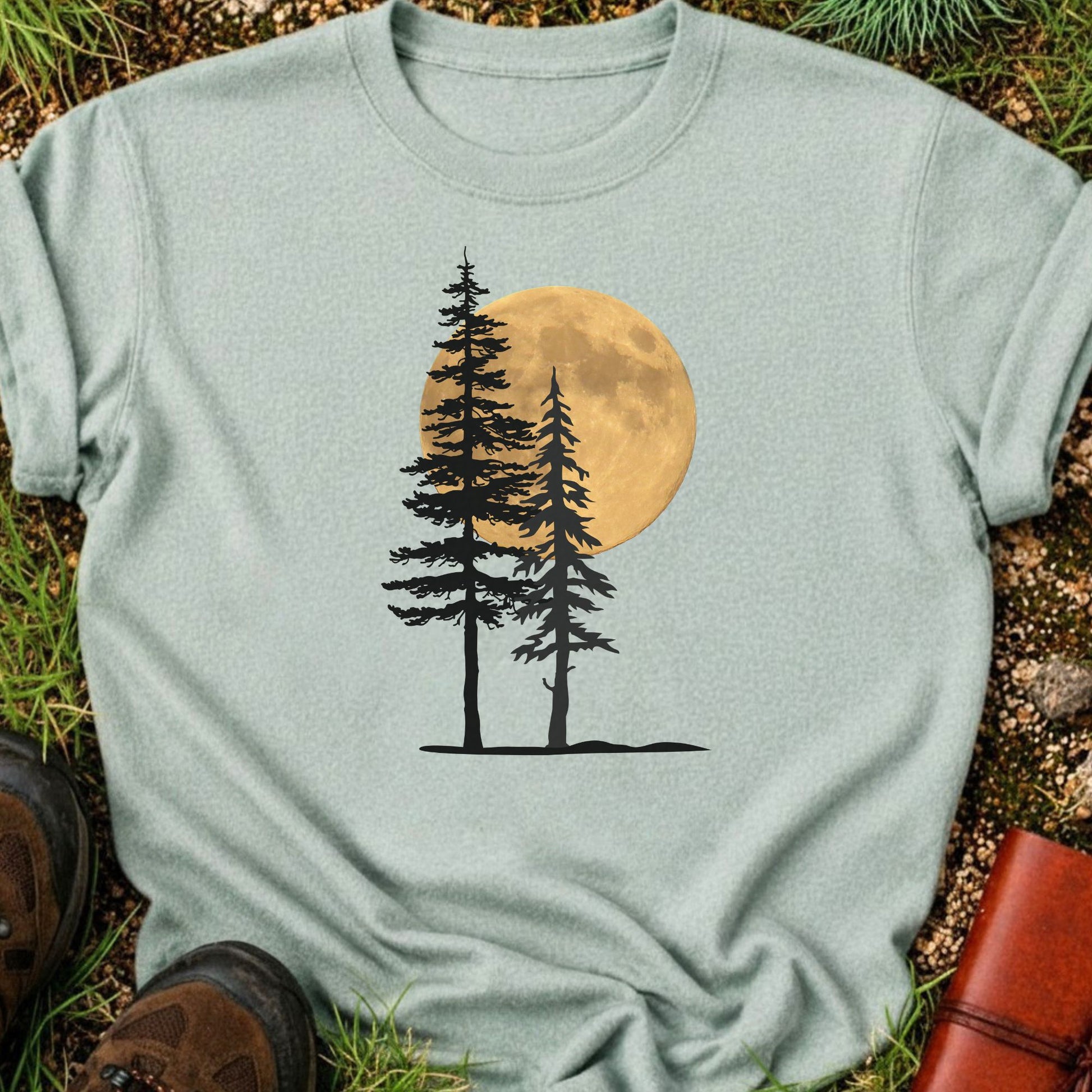 Light Green Tshirt with a graphic of  silhouetted pine trees against a glowing full moon. The Shirt is lying flat on a grassy surface.