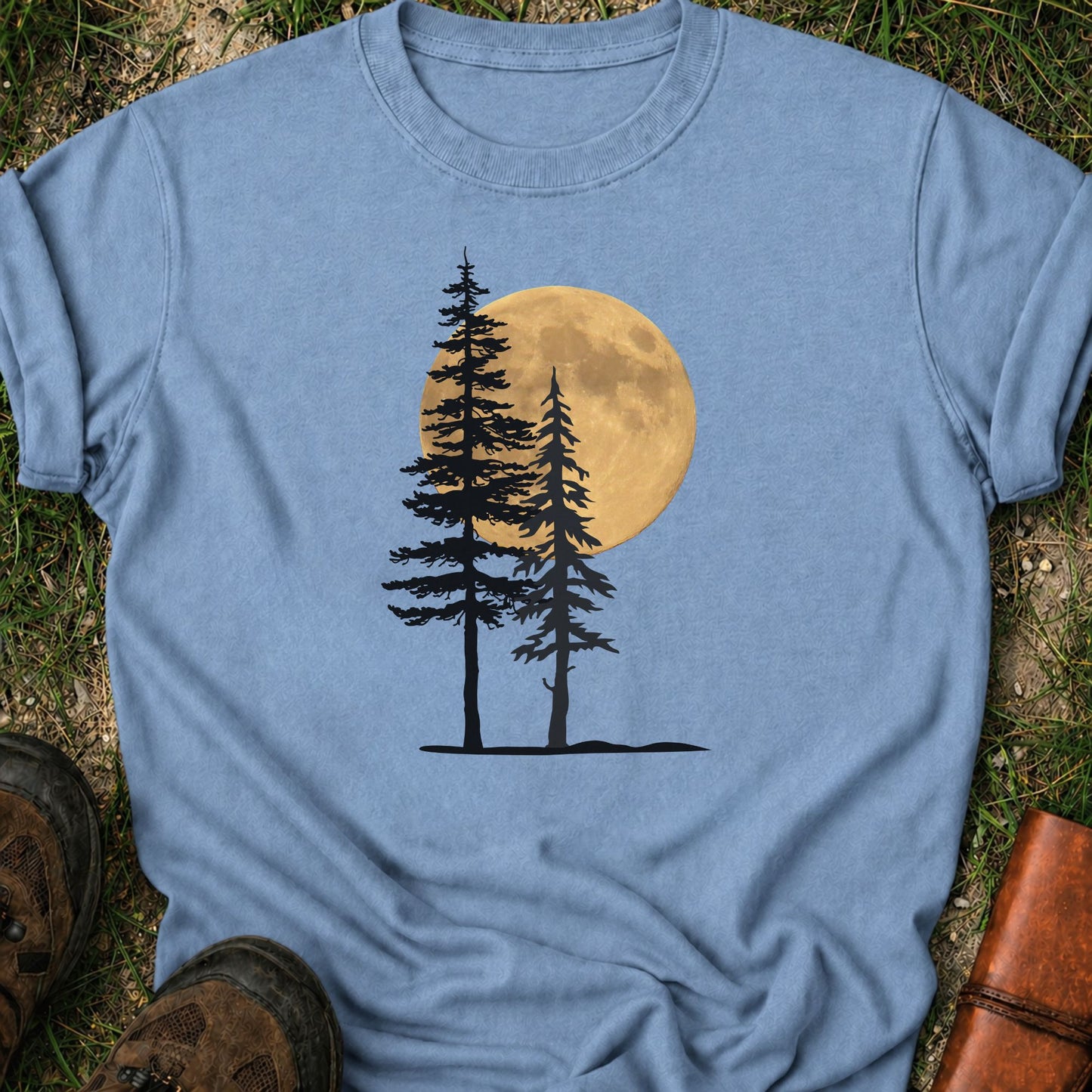 Light blue Tshirt with a graphic of  silhouetted pine trees against a glowing full moon. The Shirt is lying flat on a grassy surface.