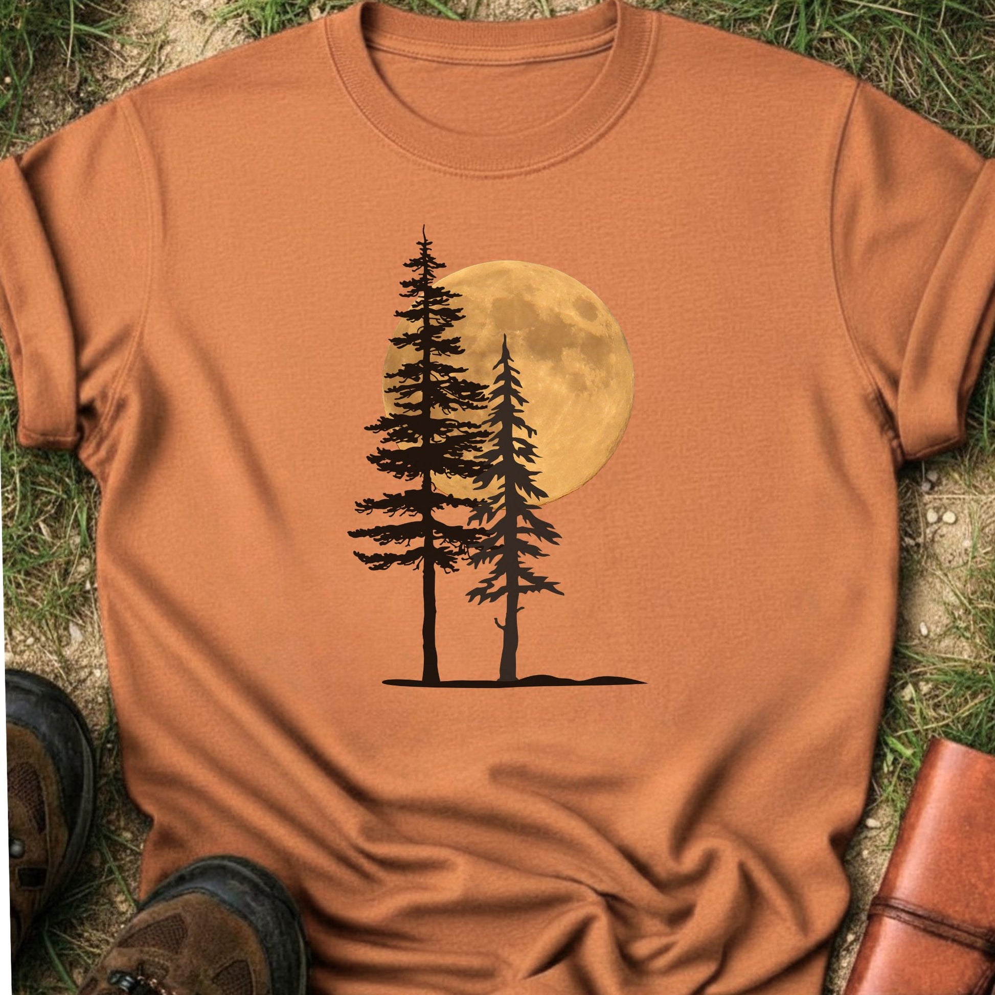 Orange Tshirt with a graphic of  silhouetted pine trees against a glowing full moon. The Shirt is lying flat on a grassy surface.