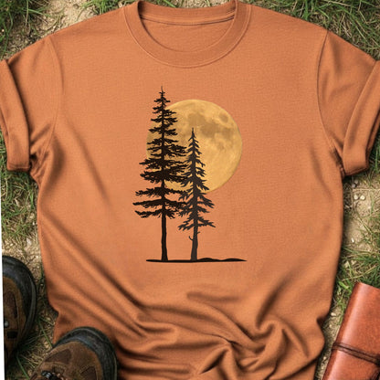 Orange Tshirt with a graphic of  silhouetted pine trees against a glowing full moon. The Shirt is lying flat on a grassy surface.