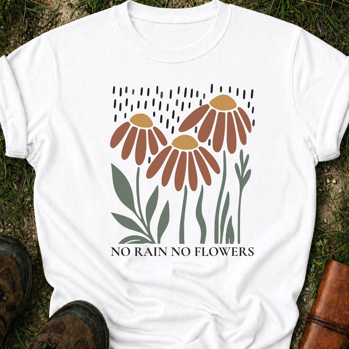 A white T-shirt featuring earthy red and gold blooms with muted green stems with text reading No Rain, No Flowers. It is lying flat on a grassy surface.