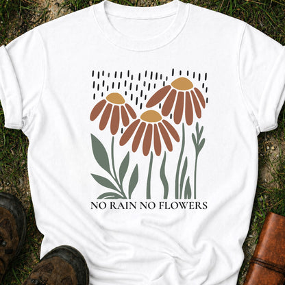 A white T-shirt featuring earthy red and gold blooms with muted green stems with text reading No Rain, No Flowers. It is lying flat on a grassy surface.