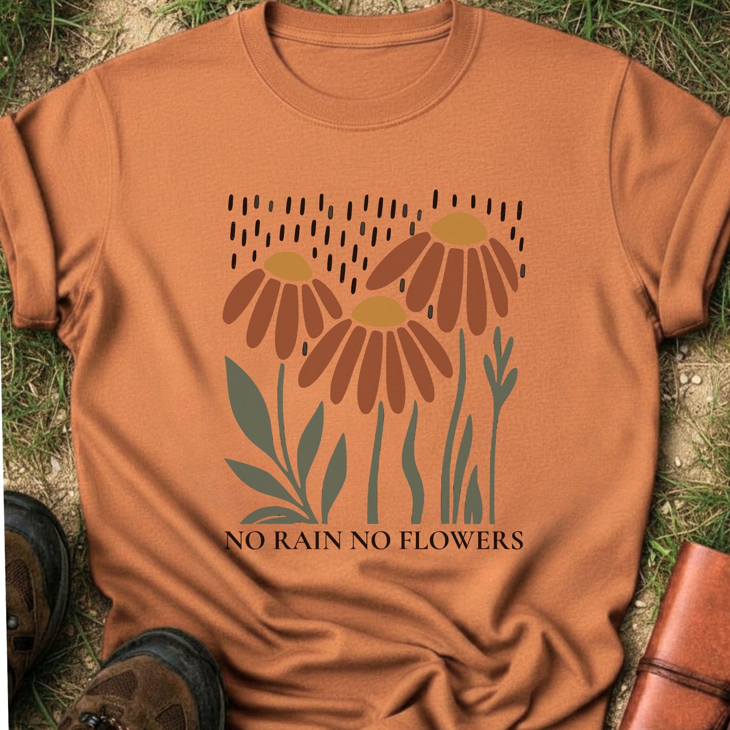 Orange T-shirt featuring earthy red and gold blooms with muted green stems with text reading No Rain, No Flowers. It is lying flat on a grassy surface.