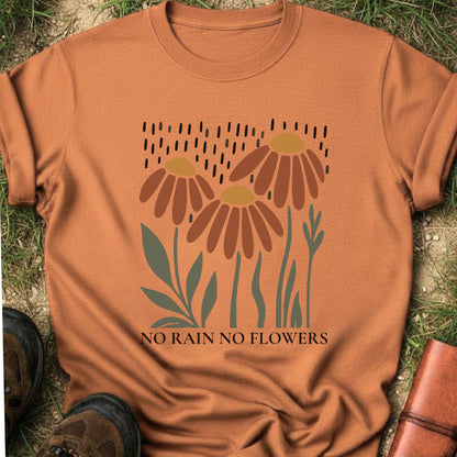 Orange T-shirt featuring earthy red and gold blooms with muted green stems with text reading No Rain, No Flowers. It is lying flat on a grassy surface.