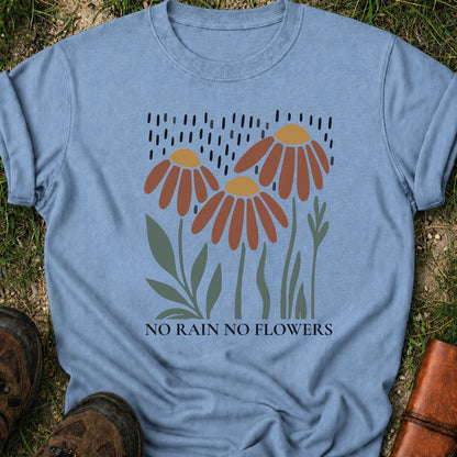 Light Blue T-shirt featuring earthy red and gold blooms with muted green stems with text reading No Rain, No Flowers. It is lying flat on a grassy surface.