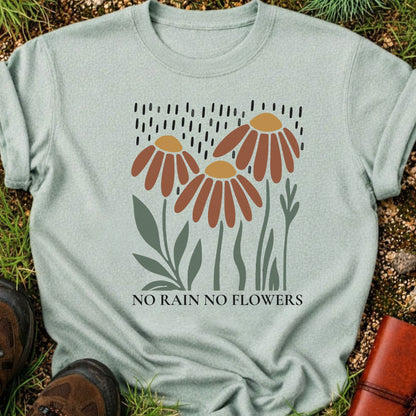 Light Green T-shirt featuring earthy red and gold blooms with muted green stems with text reading No Rain, No Flowers. It is lying flat on a grassy surface.