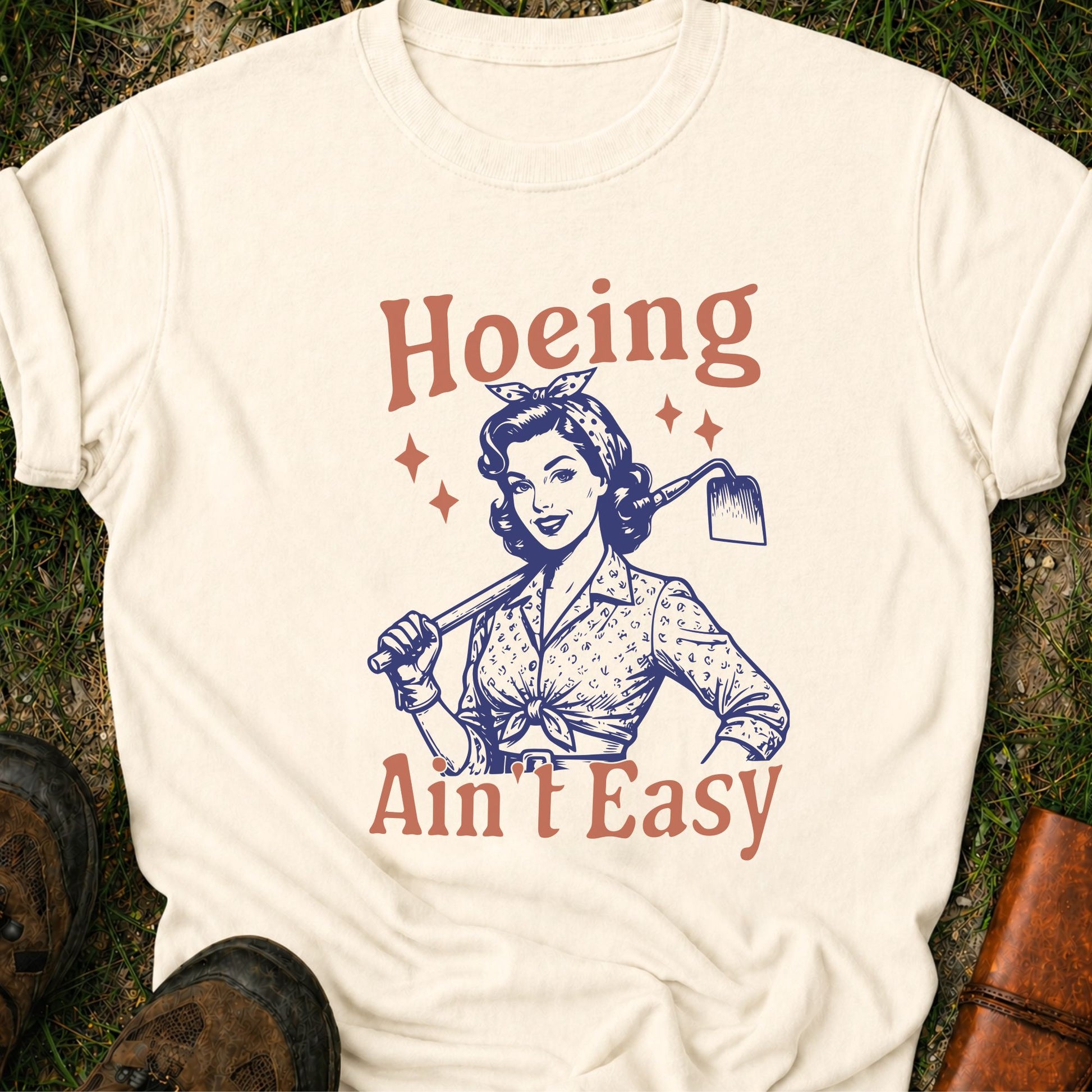 Ivory  t-shirt lying flat on a grassy surface with graphic design of a woman holding a hoe and text 'Hoeing Ain't Easy' 