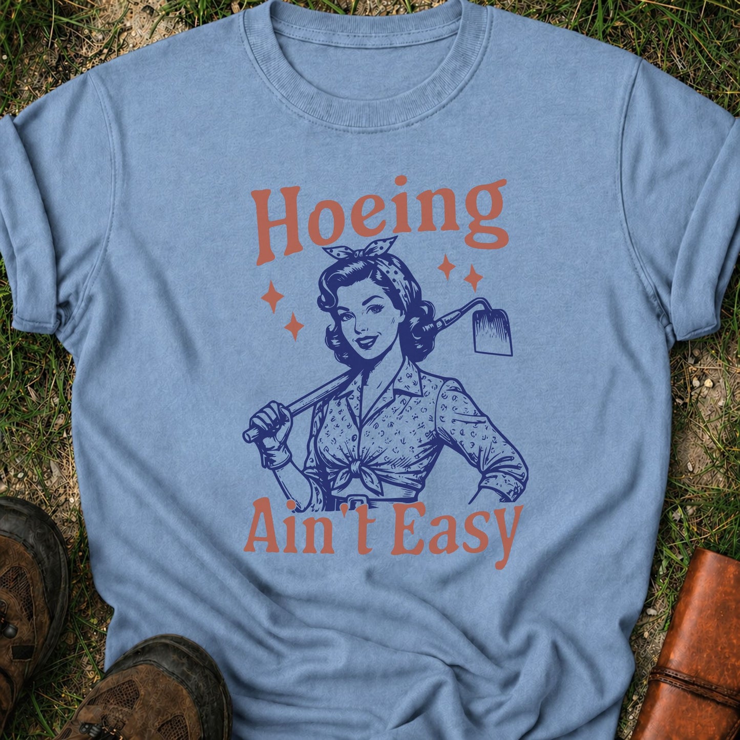 Light Blue  t-shirt lying flat on a grassy surface with graphic design of a woman holding a hoe and text 'Hoeing Ain't Easy' 