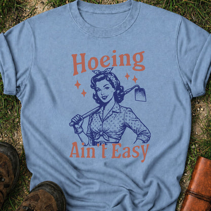 Light Blue  t-shirt lying flat on a grassy surface with graphic design of a woman holding a hoe and text 'Hoeing Ain't Easy' 