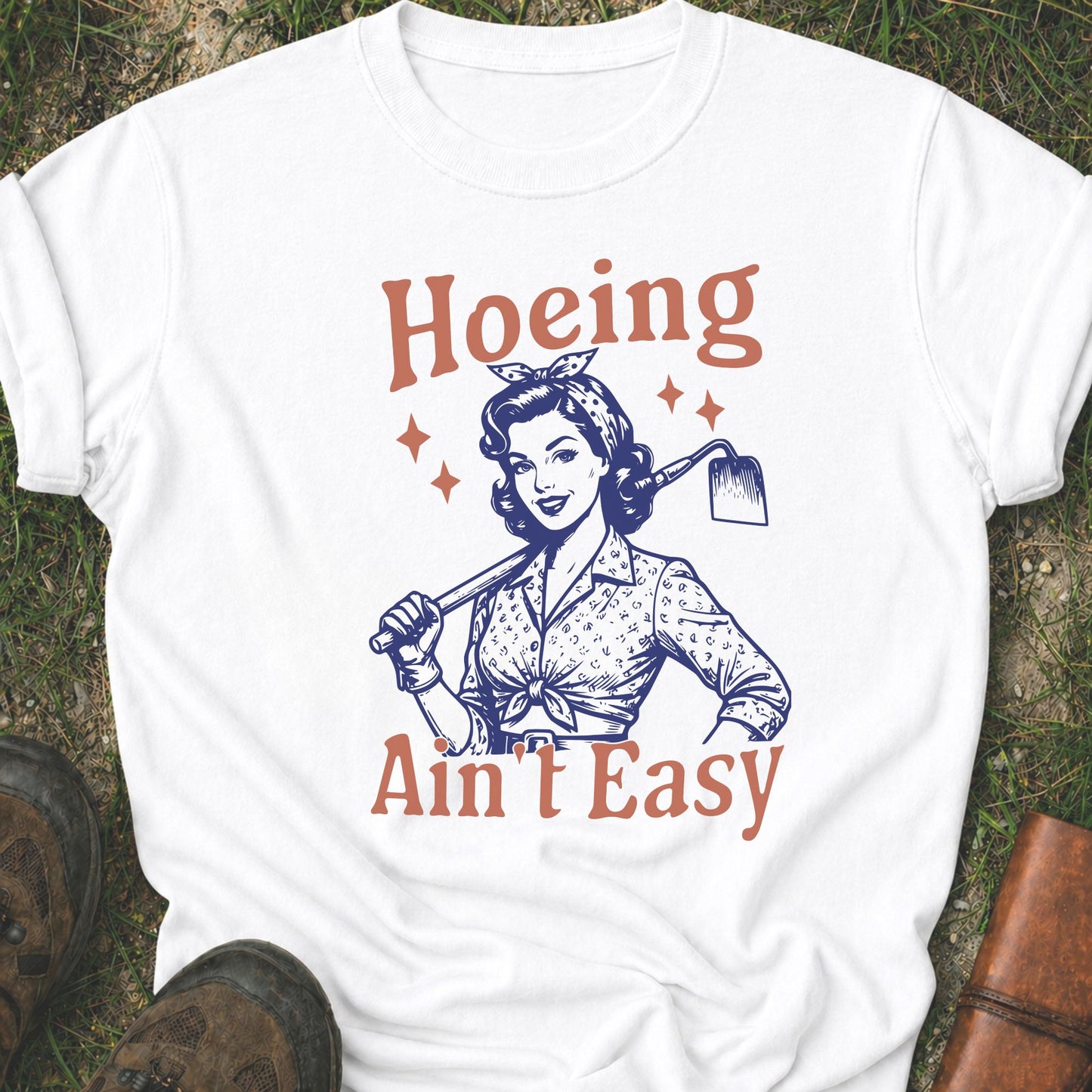 White t-shirt lying flat on a grassy surface with graphic design of a woman holding a hoe and text 'Hoeing Ain't Easy' 