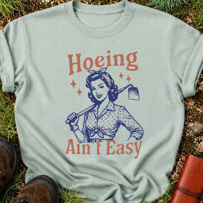  Light Green t-shirt lying flat on a grassy surface with graphic design of a woman holding a hoe and text 'Hoeing Ain't Easy' 