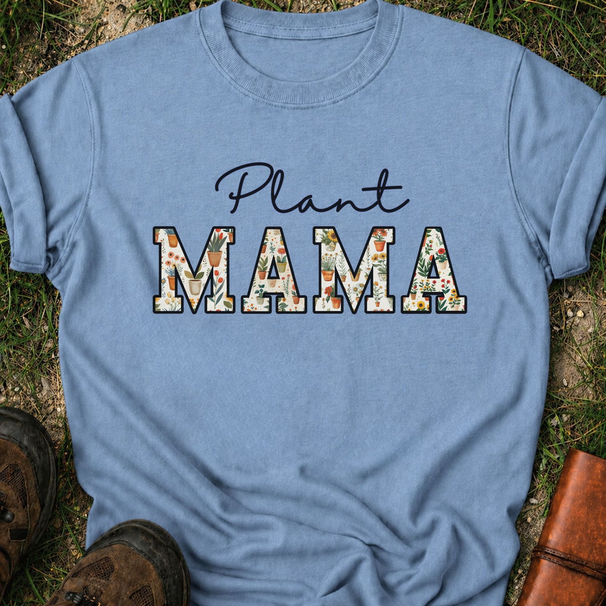 Bluet-shirt with 'Plant MAMA' text filled with cheerful potted plants and florals within the letters.  The tee is lying flat on grass
