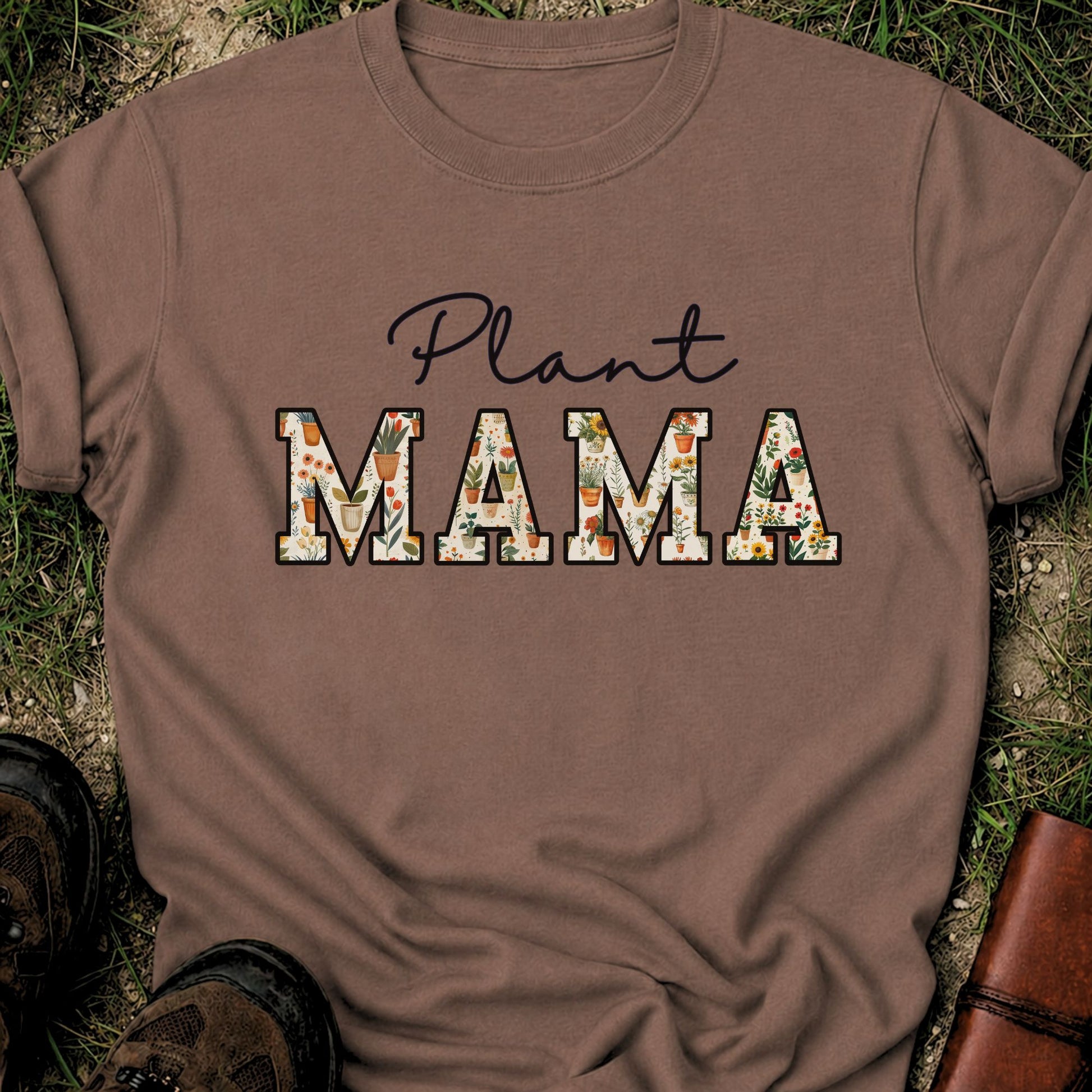 Brown t-shirt with 'Plant MAMA' text filled with cheerful potted plants and florals within the letters.  The tee is lying flat on grass