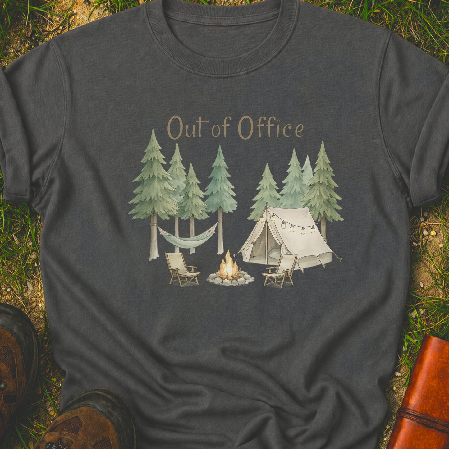 Dark Grey t-shirt with camping graphic featuring pine trees, a tent, hammock, campfire, and the phrase “Out of Office.” lying  on a grass surface