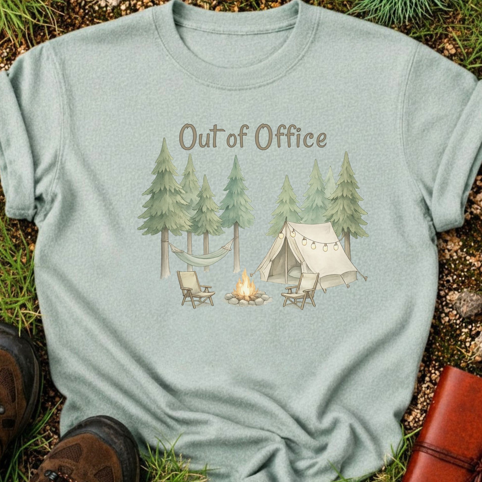 Light Green t-shirt with camping graphic featuring pine trees, a tent, hammock, campfire, and the phrase “Out of Office.” lying  on a grass surface