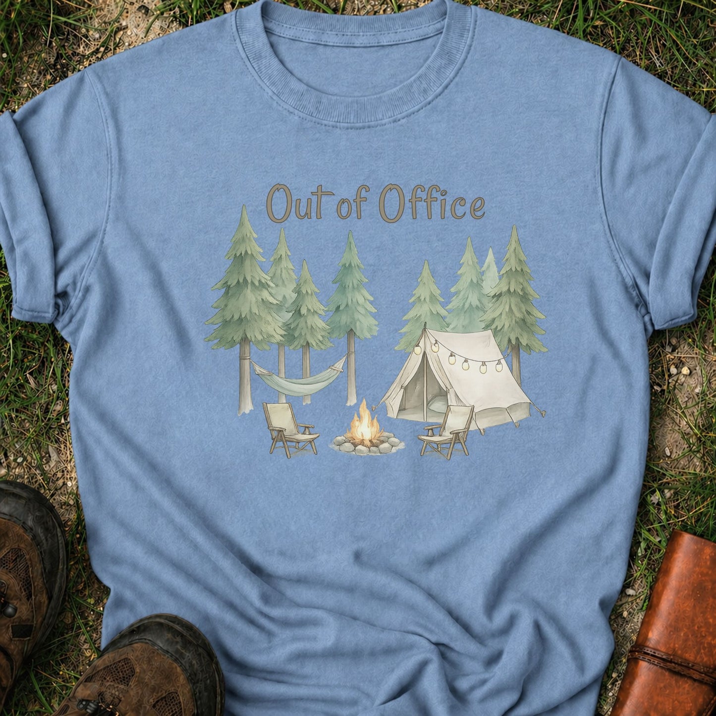 Blue t-shirt with camping graphic featuring pine trees, a tent, hammock, campfire, and the phrase “Out of Office.” lying  on a grass surface