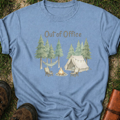 Blue t-shirt with camping graphic featuring pine trees, a tent, hammock, campfire, and the phrase “Out of Office.” lying  on a grass surface