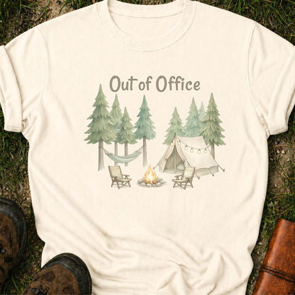 Ivory t-shirt with camping graphic featuring pine trees, a tent, hammock, campfire, and the phrase “Out of Office.” lying  on a grass surface