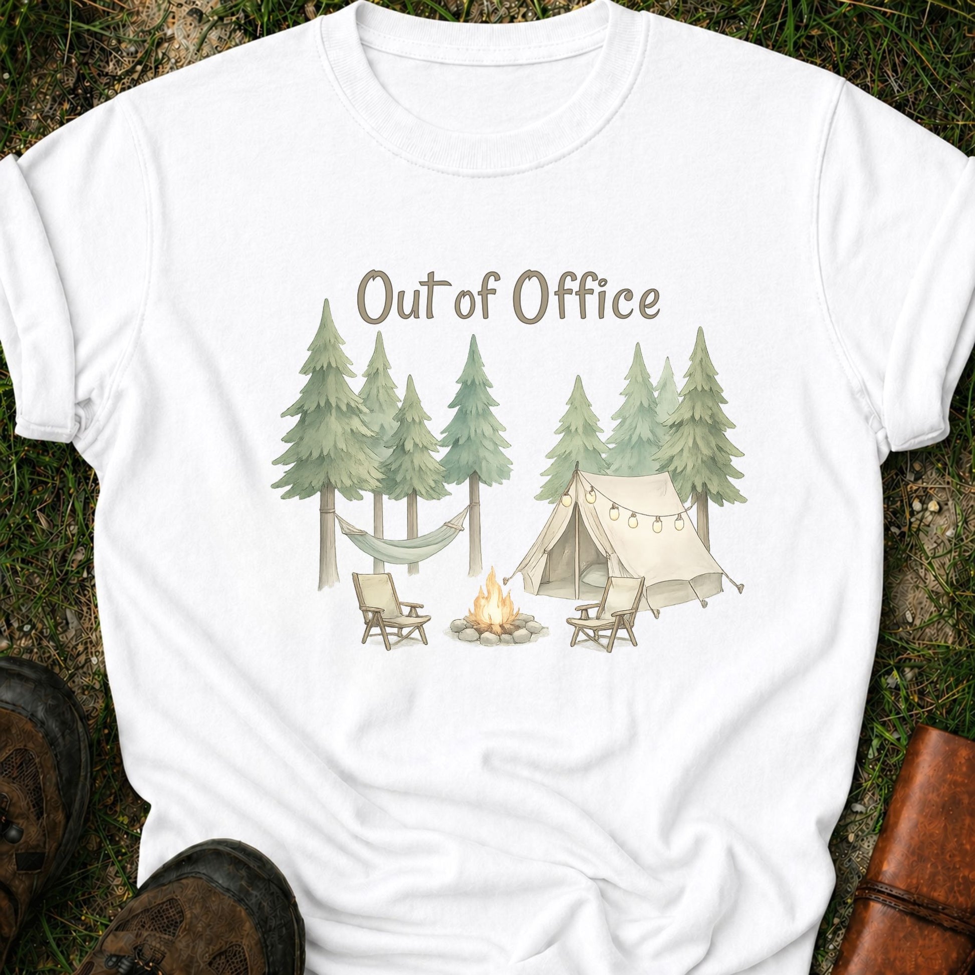 White t-shirt with camping graphic featuring pine trees, a tent, hammock, campfire, and the phrase “Out of Office.” lying  on a grass surface.