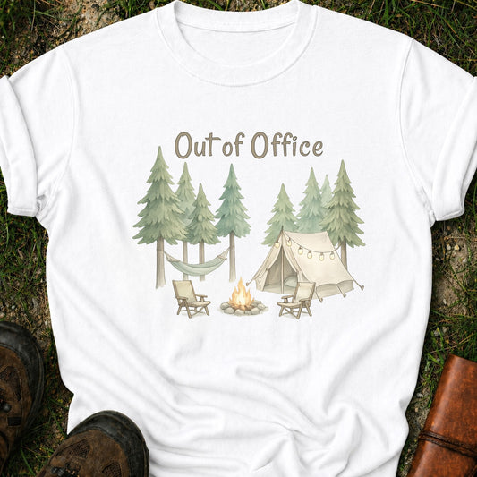 White t-shirt with camping graphic featuring pine trees, a tent, hammock, campfire, and the phrase “Out of Office.” lying  on a grass surface.