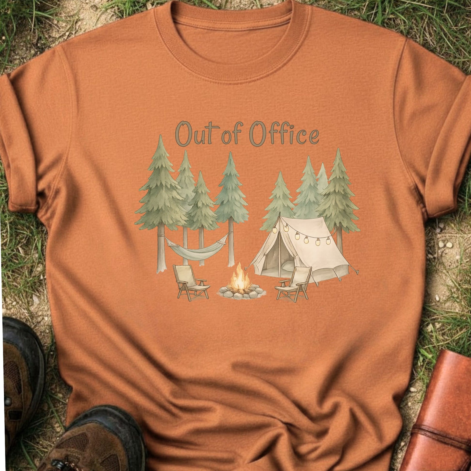 Orange t-shirt with camping graphic featuring pine trees, a tent, hammock, campfire, and the phrase “Out of Office.” lying  on a grass surface