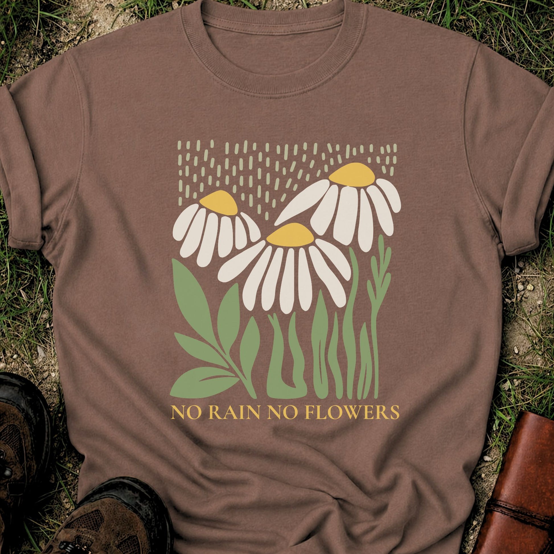 Brown T-Shirt with 3 boho style flowers in the rain with the text "No Rain No Flowers".  Tshirt is lying flat on grass.