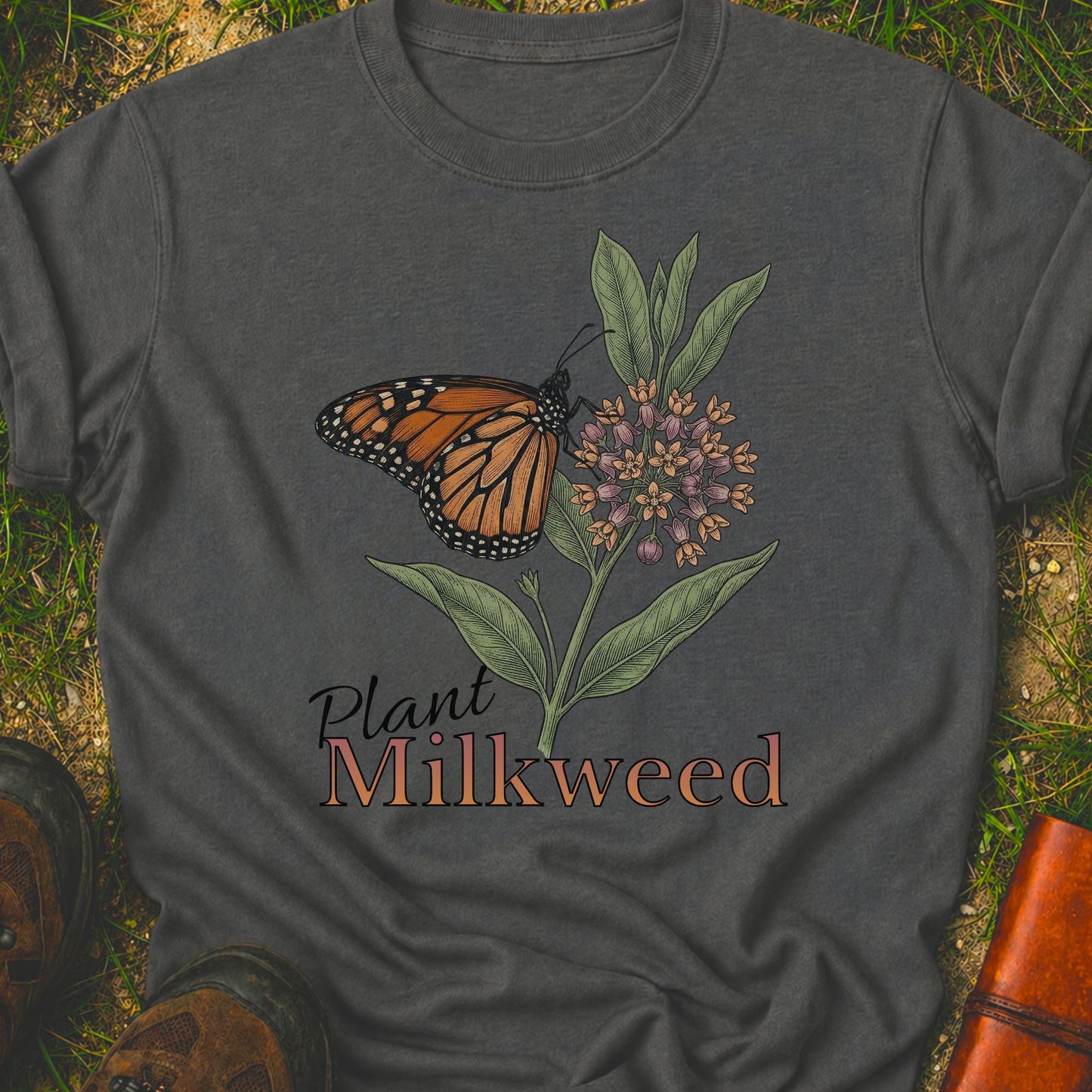 Dark gray t-shirt with a butterfly and milkweed design  and the caption Plant Milkweed lying  on the grass