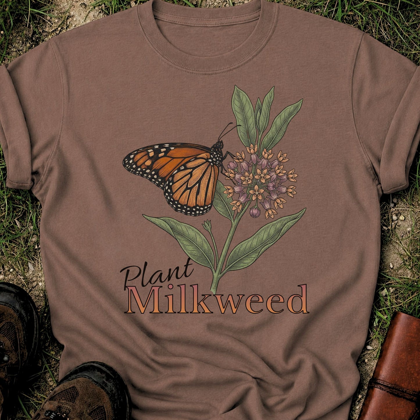 Brown t-shirt with a butterfly and milkweed design  and the caption Plant Milkweed lying  on the grass