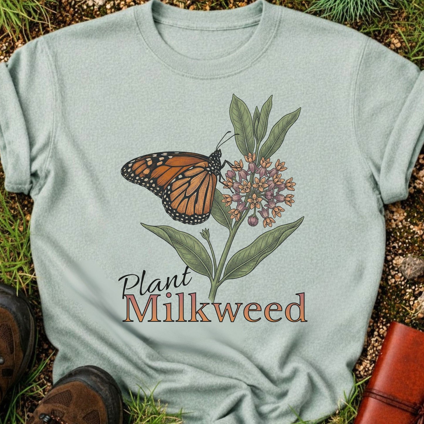 LIght Green t-shirt with a butterfly and milkweed design  and the caption Plant Milkweed lying  on the grass