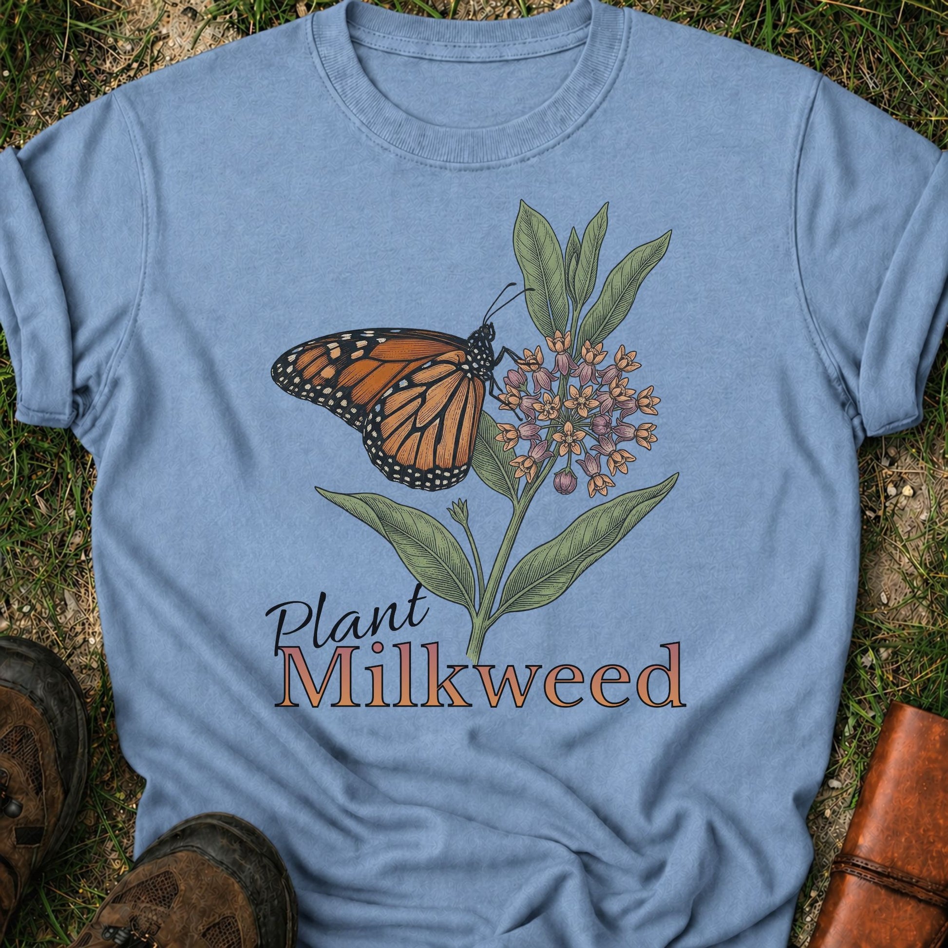 Blue t-shirt with a butterfly and milkweed design  and the caption Plant Milkweed lying  on the grass