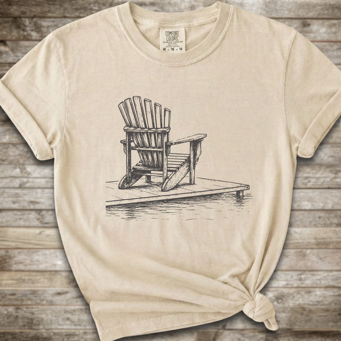 An Ivory short-sleeve t-shirt with a white line art graphic of a Muskoka / Adirondack chair on a dock, styled with a knot at the bottom hem and laid flat on a rustic wood 