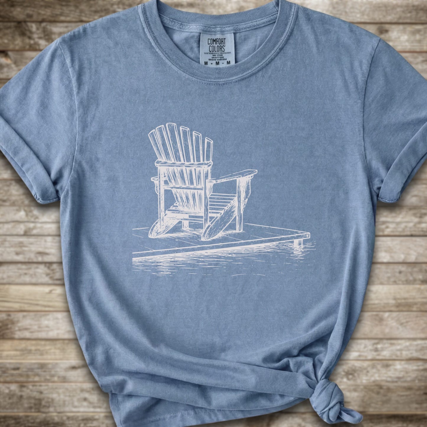 A heathered blue short-sleeve t-shirt with a white line art graphic of a Muskoka / Adirondack chair on a dock, styled with a knot at the bottom hem and laid flat on a rustic wood background.