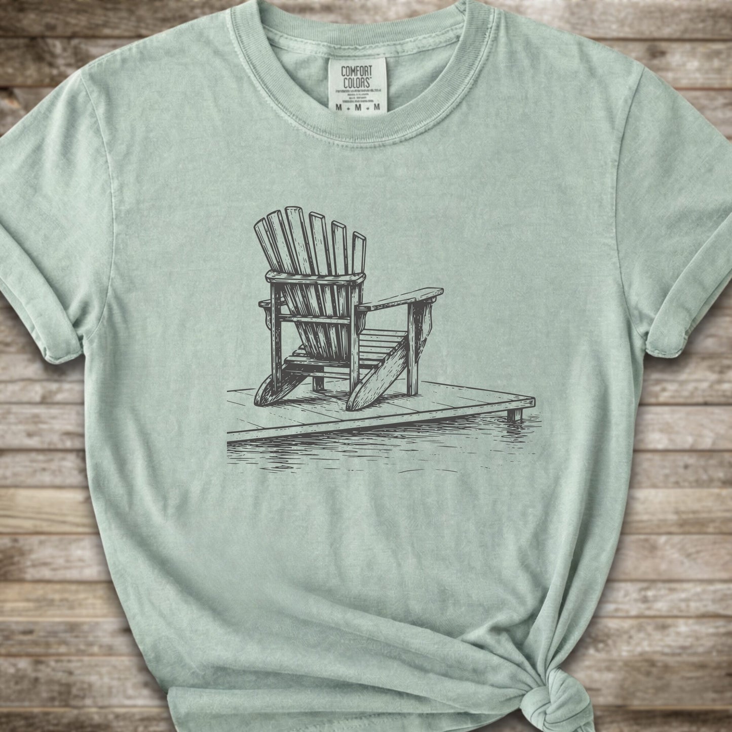 A light green short-sleeve t-shirt with a white line art graphic of a Muskoka / Adirondack chair on a dock, styled with a knot at the bottom hem and laid flat on a rustic wood 