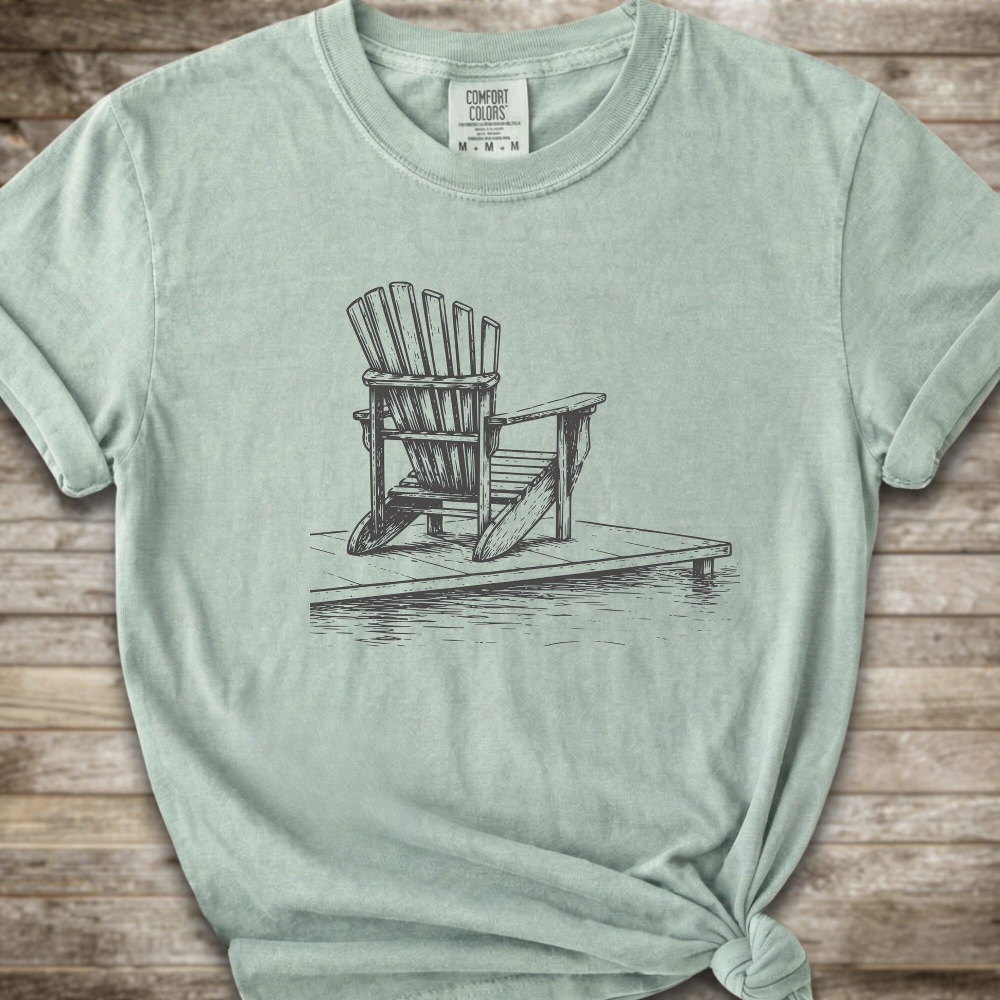 A light green short-sleeve t-shirt with a white line art graphic of a Muskoka / Adirondack chair on a dock, styled with a knot at the bottom hem and laid flat on a rustic wood 