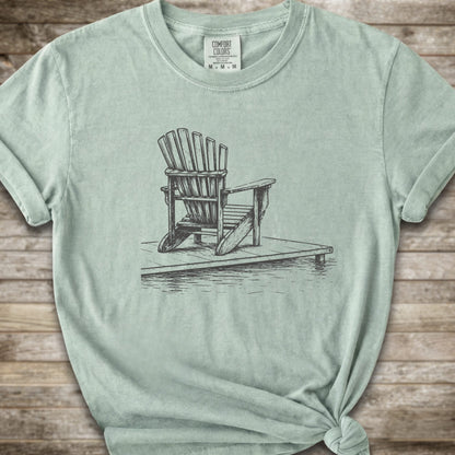A light green short-sleeve t-shirt with a white line art graphic of a Muskoka / Adirondack chair on a dock, styled with a knot at the bottom hem and laid flat on a rustic wood 