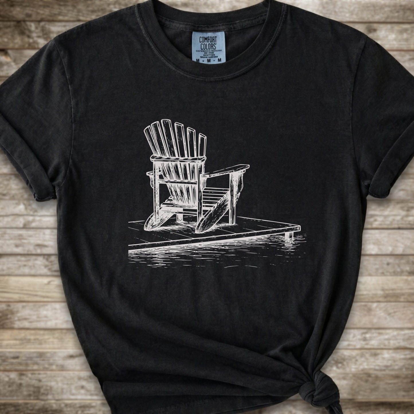 A black short-sleeve t-shirt with a white line art graphic of a Muskoka / Adirondack chair on a dock, styled with a knot at the bottom hem and laid flat on a rustic wood 