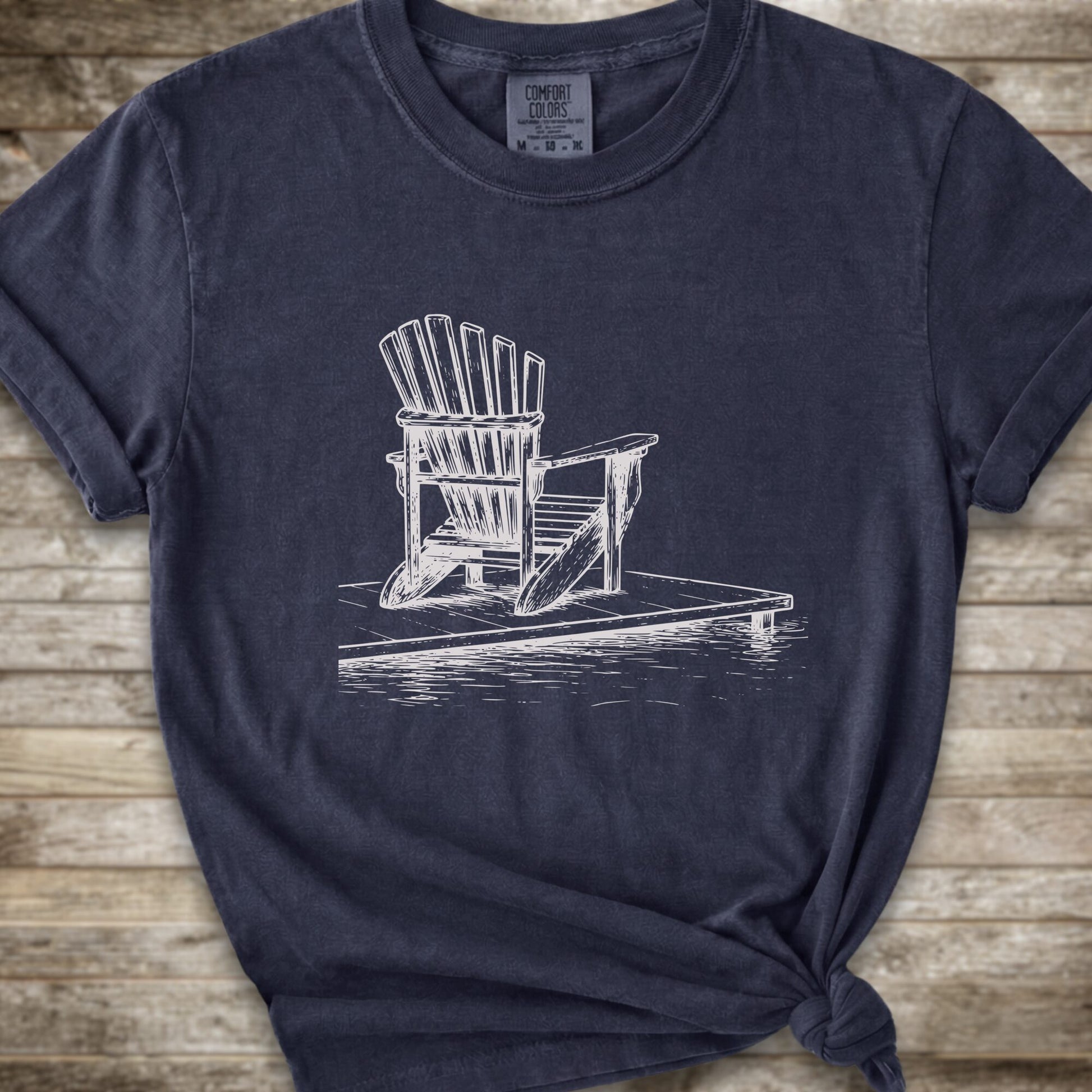 A lnavy blue short-sleeve t-shirt with a white line art graphic of a Muskoka / Adirondack chair on a dock, styled with a knot at the bottom hem and laid flat on a rustic wood 
