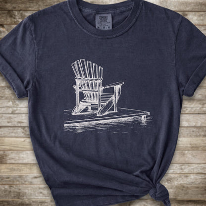 A lnavy blue short-sleeve t-shirt with a white line art graphic of a Muskoka / Adirondack chair on a dock, styled with a knot at the bottom hem and laid flat on a rustic wood 